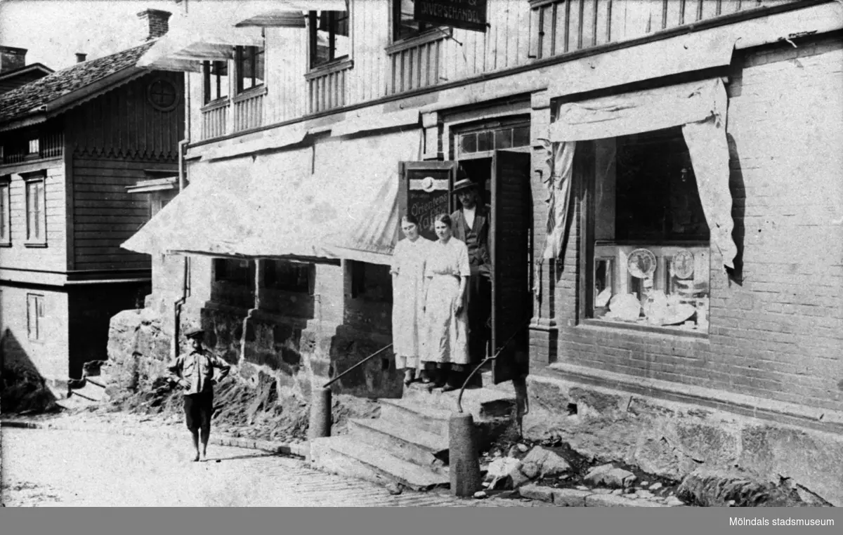 Örns affär vid "Kråkan" i Mölndal på 1910-talet. Vid ingången, på trappan står två kvinnor (troligen butiksbiträden) och en man. Nedanför står en pojke.
