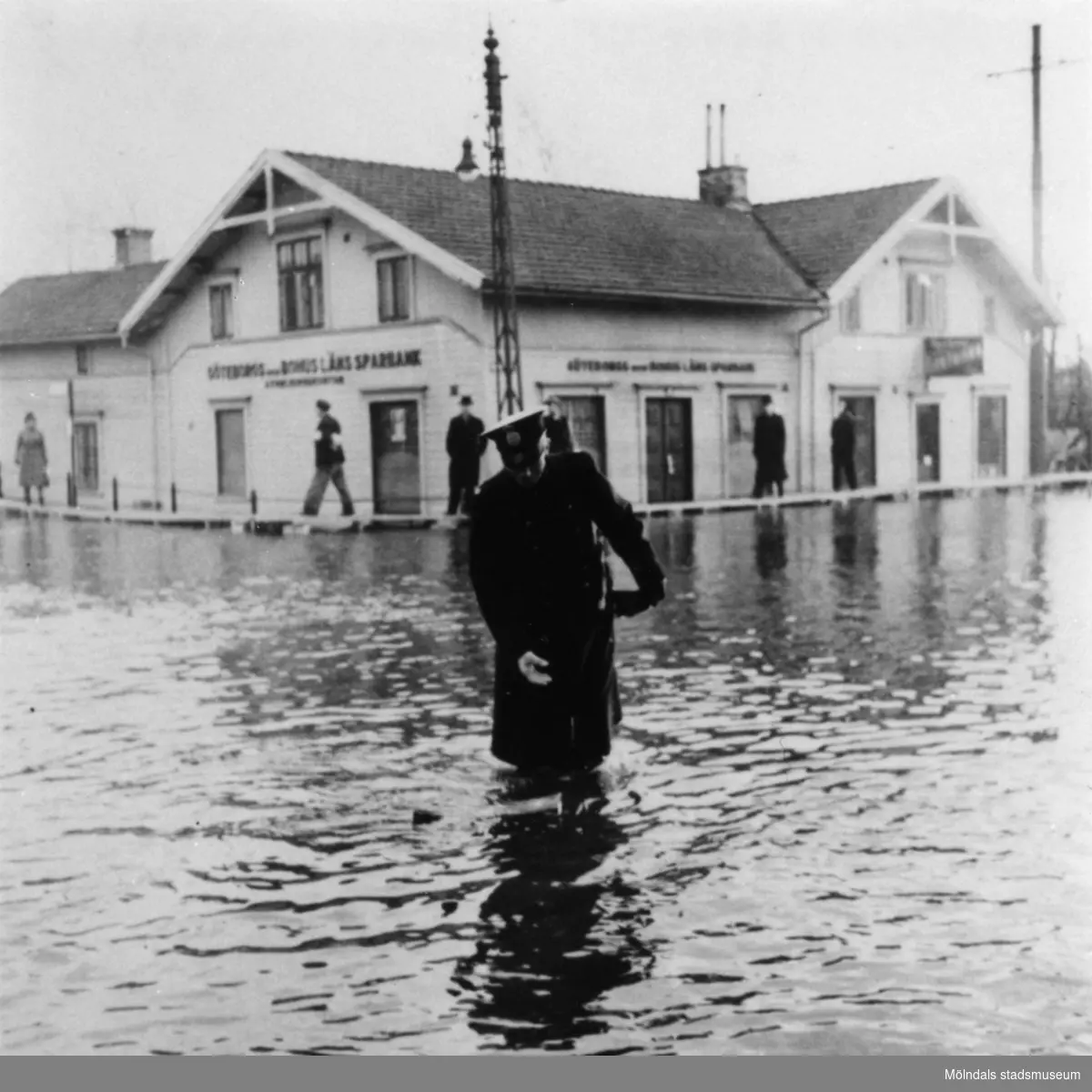 Uniformsklädd man (Polisman) under tjänstgöring vid översvämning på Göteborgsvägen i Mölndalsbro. Göteborgs- och Bohusläns sparbank syns i bakgrunden. Omkring 1920