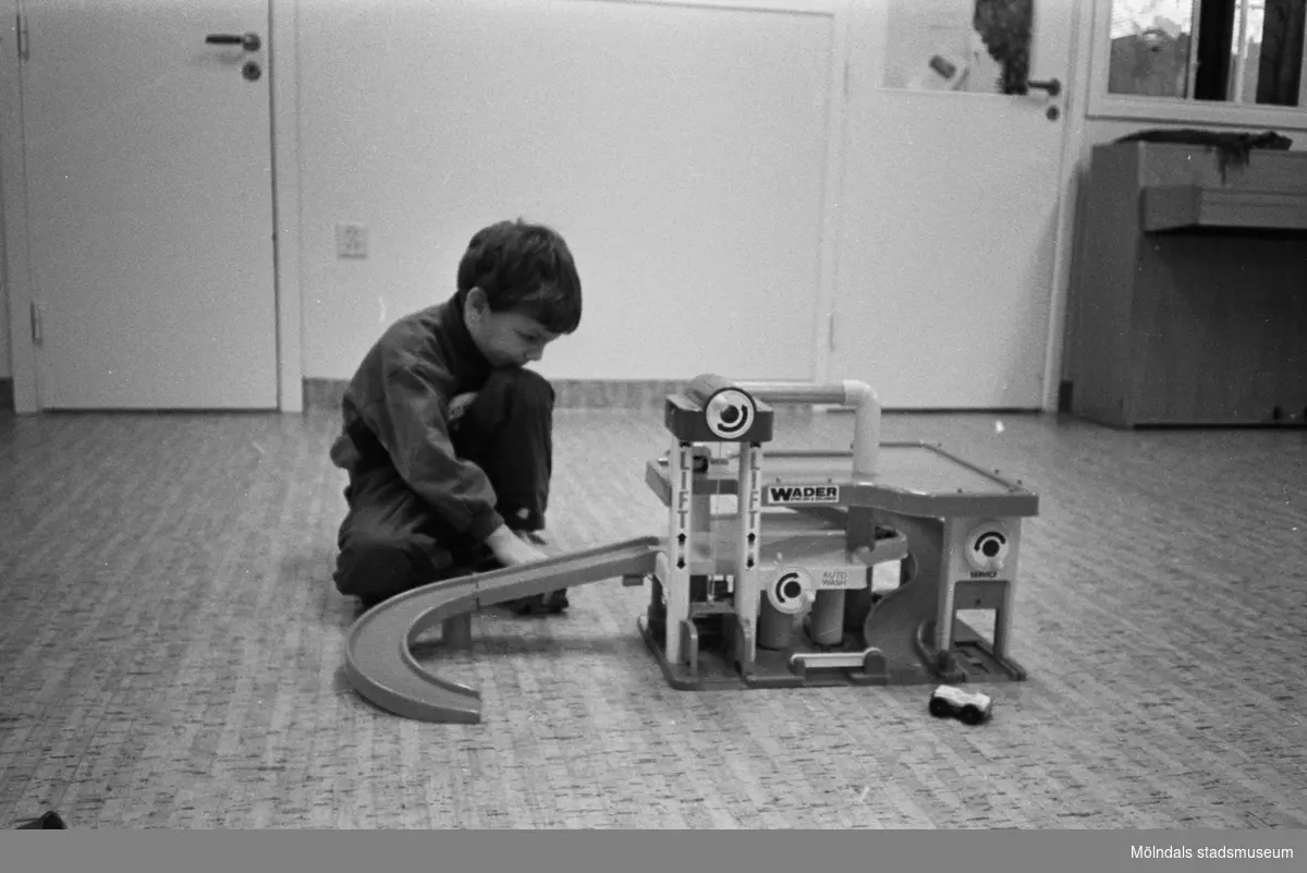 En pojke sitter och leker med ett Wader-garage utrustat med bensinpumpar, hiss, tvätt- och servicestation samt en lång avfart för tillhörande leksaksbilar. Han sitter på ett korkliknande linolium-golv. I bakgrunden till vänster syns en stängt dörr. Till höger står ett piano.