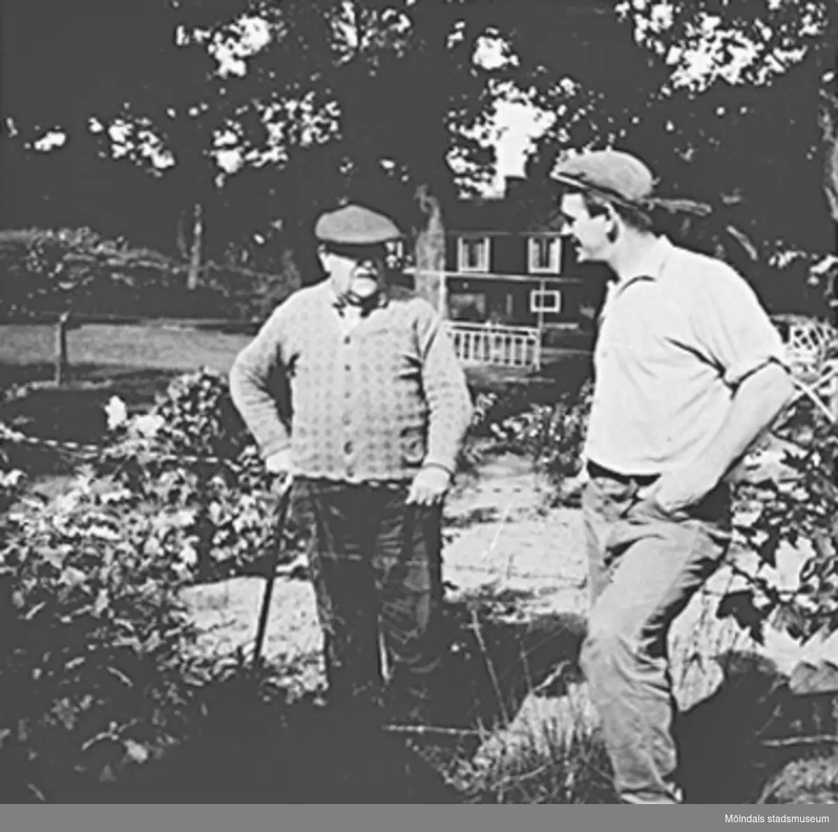 Två män står i en trädgård i Kållered, 1940-tal. I bakgrunden ses en stuga.