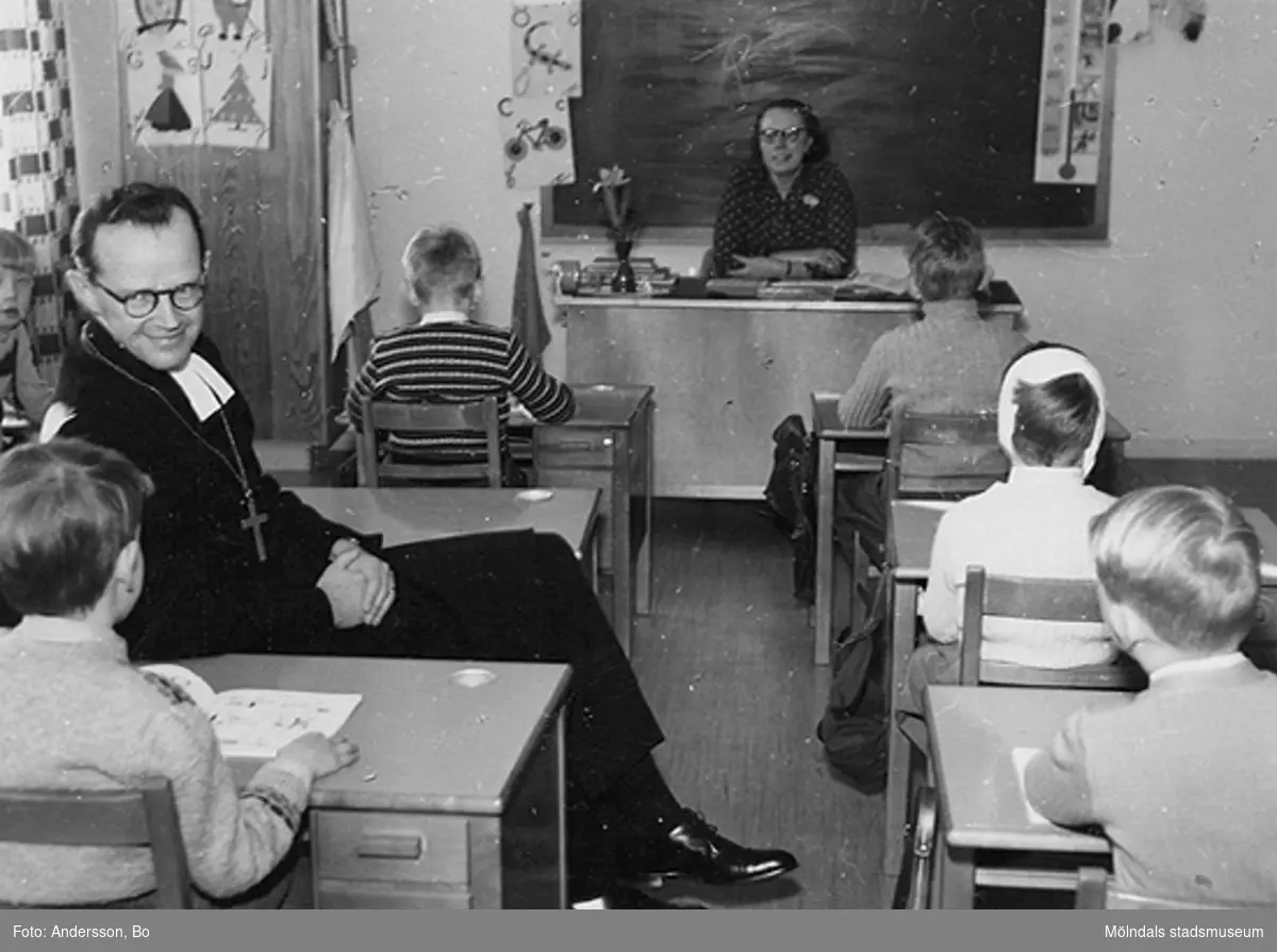 Biskop Bo Giertz besöker Brattåsskolan och sitter bland eleverna i ett klassrum 14 feb 1955. Vid katedern sitter Lärarinnan Greta Andersson (född Alm).