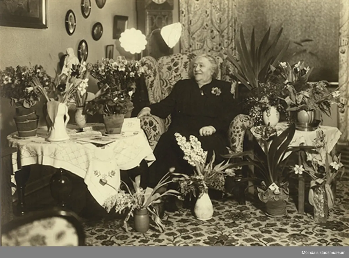 En äldre kvinna i ett hem, omgiven av blommor, 1930-tal. Antagligen firar hon sin födelsdag.