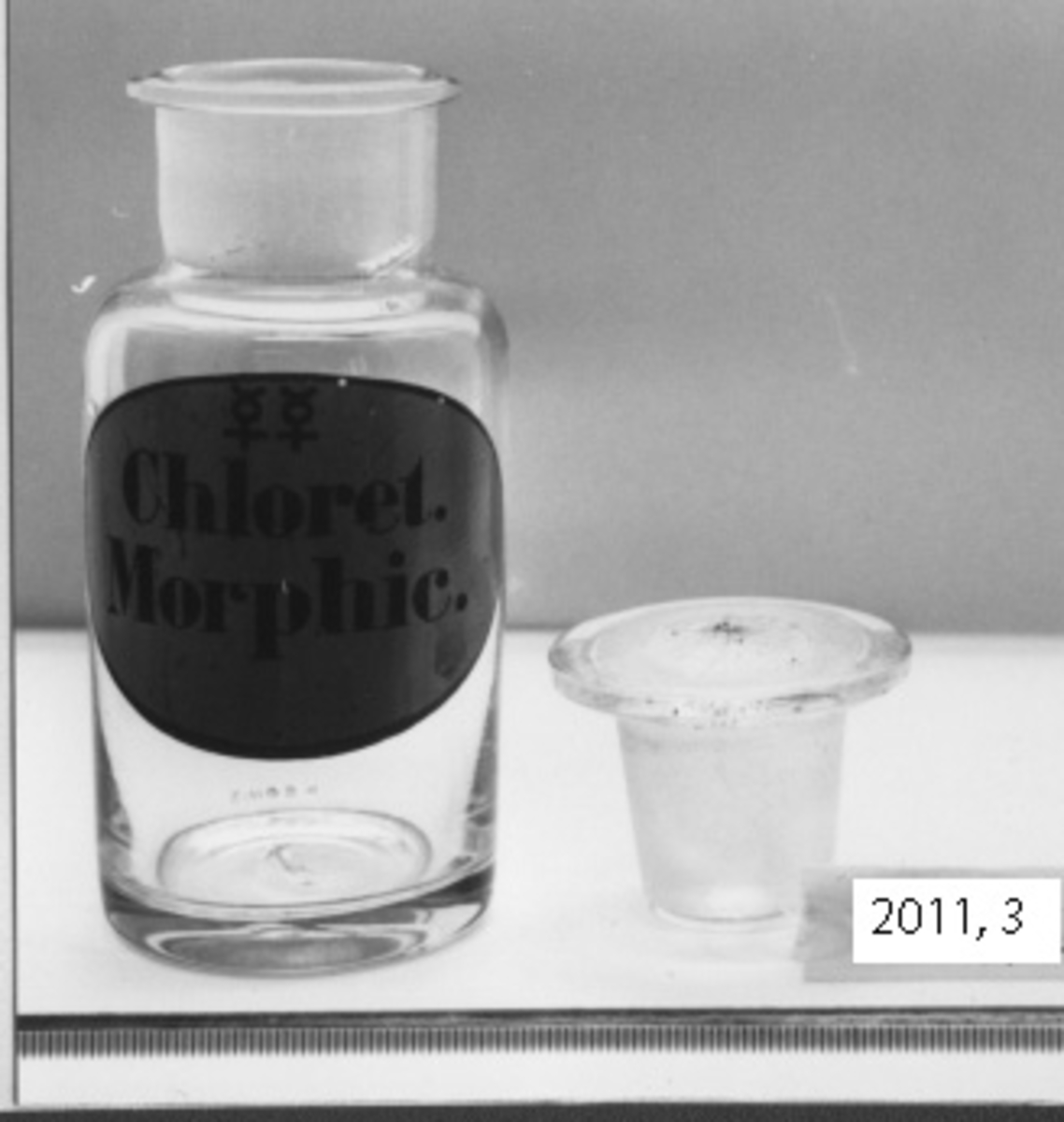 Flaska av klart glas för medicin. Den är cylindrisk och försedd med glaspropp med runt, kupigt huvud. På sidan är målad i blått ovalformad etikett med kant i svart. Märkning i svart (se bild) Chloret Morphic.