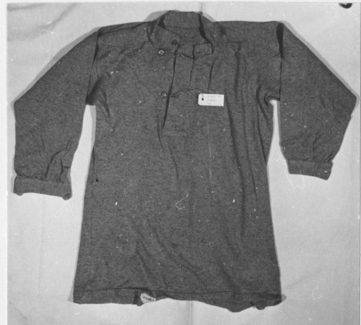 Skjorta av grått bomullstyg. Knäpps famtill och runt halsen med tre knappar av bleck och runt handloven med en knapp.
Märkning: 1 y 263 205 samt 3 kronstämplar, i svart.
