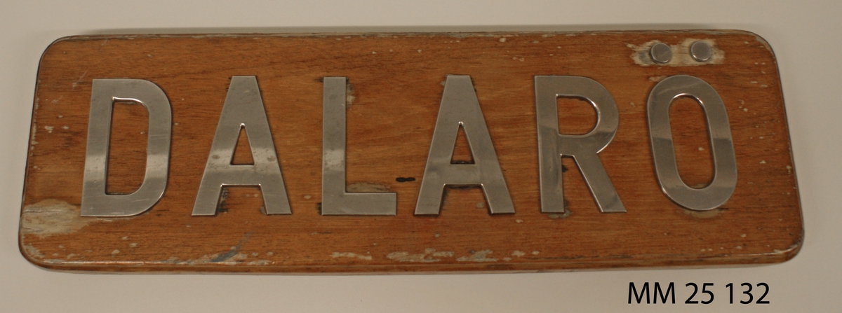 Namnbräda till vedettbåten Dalarö. Rektangulär fernissad träplatta med bokstäver av stål bildande namnet "Dalarö". Bokstäverna skruvade i träplattan.