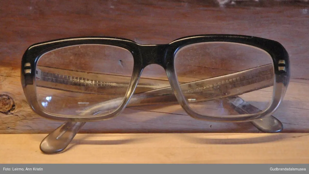 Et par briller med store glass og kraftigere innfatning i hardplast, metallet i brillestengene ses gjennom siden plasten er transparent. Brillen er svart øverst og lys grågrønn/oransje rundt på innfatningen ellers. Glass med nedre lesefelt. Brukt i reparasjonsarbeid, nærarbeid.