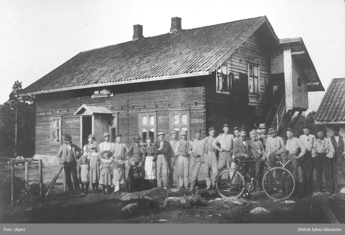 Storebrakka på Refsahl i Torsnes i Borge, for stenhoggere, ca. 1890. Alle på bildet bodde i huset, formann Olsen inklusive. Formannen står med sykkel i forgrunnen, har skalk og sløyfe. Bygningen brant i 1918-19.