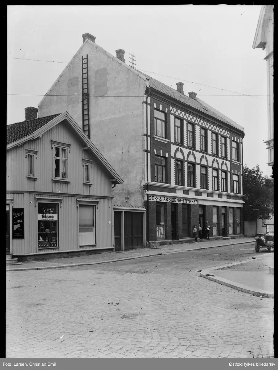 Gateparti fra Pellygata 61,  Sarpsborg - murbygning til høyre. 
Kobberstads Handelsskole holdt til der ca 1940. I bygning tll høyre var det da bildet ble tatt  også  "Bok- og aksidenstrykkeri". 

Bygning til venstre har gatenummer 63. Reklameskilt for Melange og Rinso, antakelig en kolonialforretning. Kan være innehaver  J. Sikkeland, som hadde adresse Pellygata 63 i 19143. Ant.   Johannes Jensen Sikkeland.

Bilen til høyre i bildet: 
B-3412, Ford T, eier Ragnar Mathisen, Sarpsborg, i 1928.
Bildet tatt etter 1925, før det gikk ikke bilnumrene så høyt. Forden er årsmodell 1926-27.