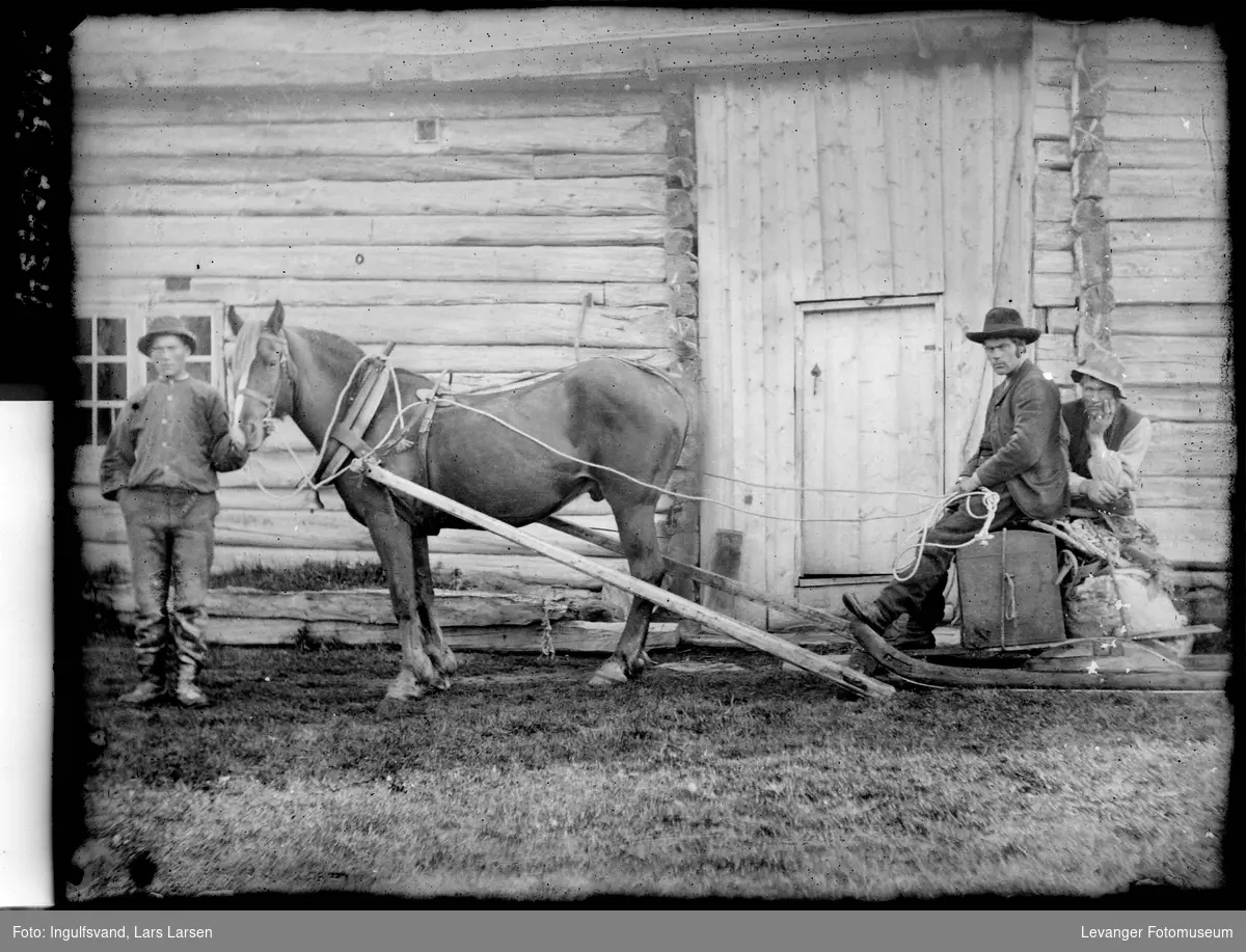 Hest på barmark med slede foran en lafta uthusbygning, to menn sitter på lasset  mens en annen mann holder hestens hodelag.