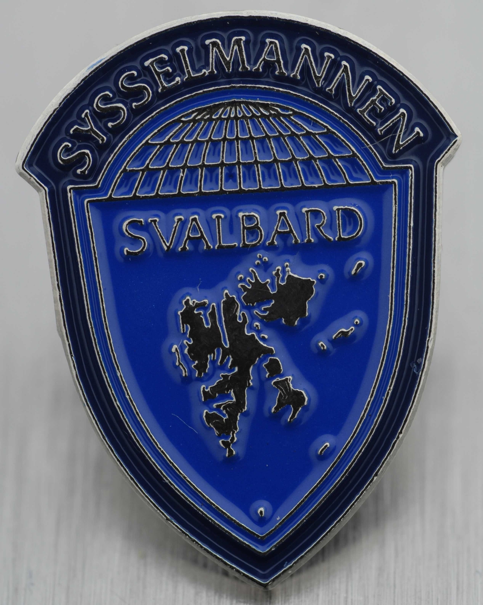 Emaljert pin med Sysselmannens logo i fargene blått og sølv. Formet som et skjold med den nordlige halvkule på toppen med teksten 'Sysselmannen' over, kartet av Svalbard under.