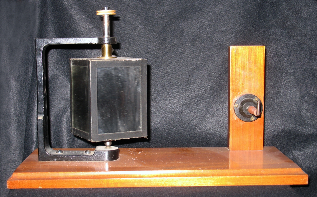 Ett mätinstrument på en bottenplatta i trä. På bottenplattan finns ett stativ med en kub vars sidor är täckta med spegelglas. Spegelkuben kan rotera.