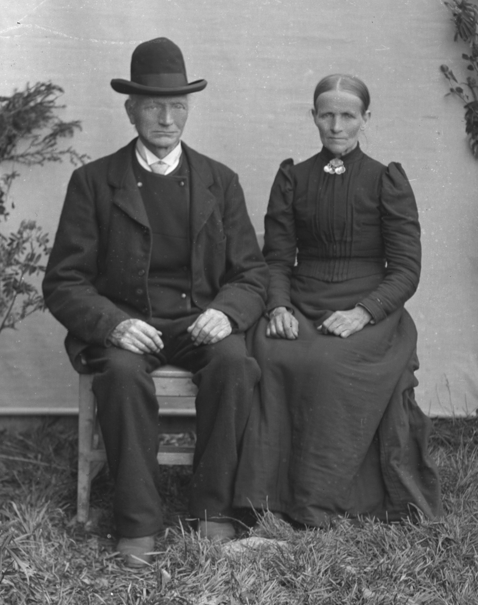 Dresskledd mann med hatt, og kvinne kledd i mørk kjole, sittende foran hvitt lerret