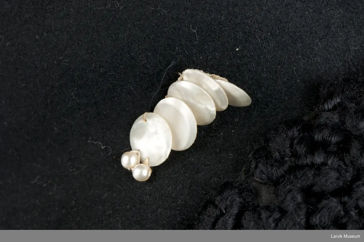 Form: sort filt, langs forsiden besetning av sort persianerimitasjon og garnityr av 5 plater perle- morsimitasjon og 2 små perler, filtsløyfe bak, sort ripsbånd på innsiden, omsydd 