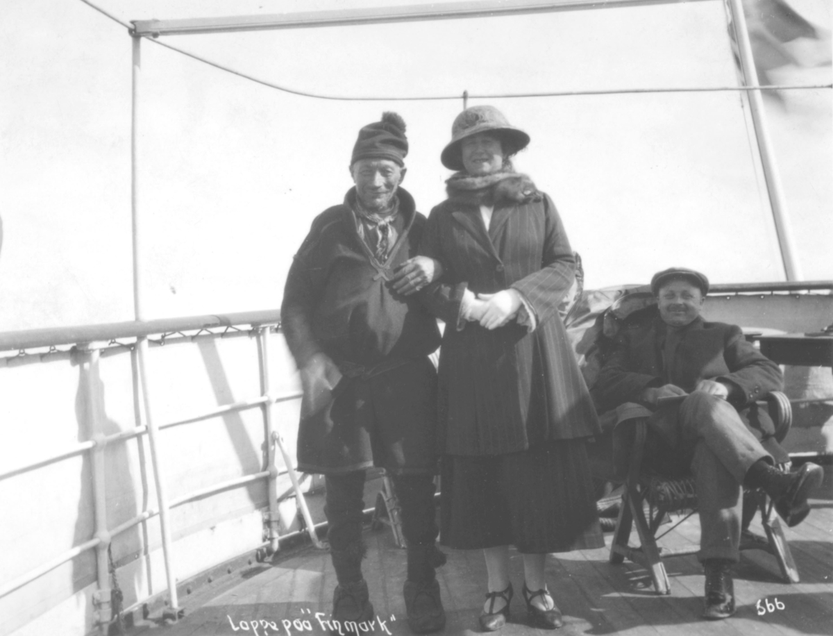 Passasjerer ombord på Hurtigruteskipet DS "Finmarken".  En mann i samisk kofte står til venstre for en kvinne i kåpe med hatt og skjerf. En mann sitter i en av dekksstolene ombord.