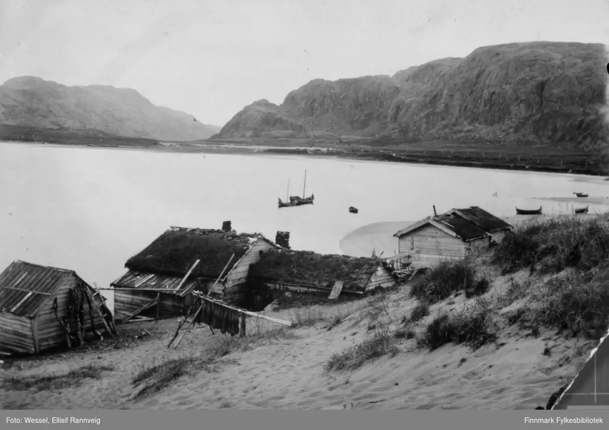 Fiskebuer på norsk side av elvemunningen i Grense Jakobselv. To seilbåter i bakgrunnen. Bak bildet står det "Fiskerhytter ved Grense Jakobselvens munding".