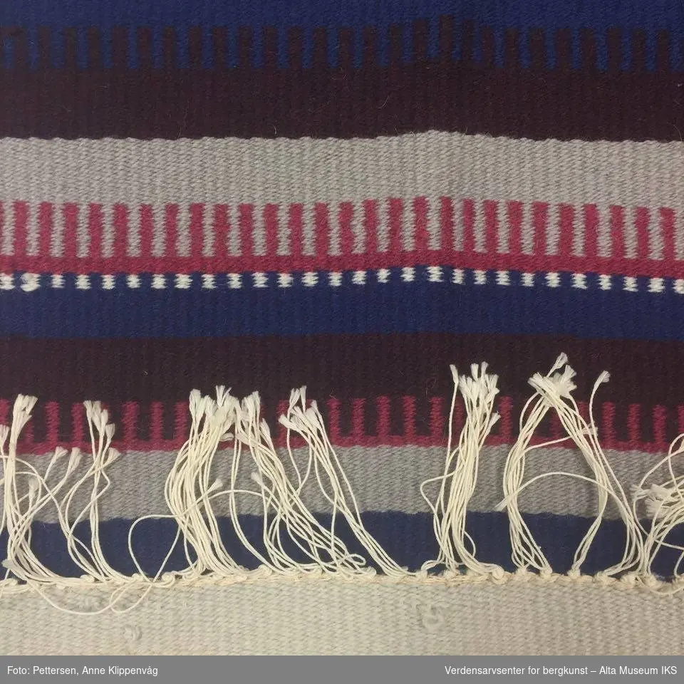 Rektangulær rye med stripete, flerfarget mønster. Rysjer av hvite tråder i hver ende. På den ene kortenden er det dekorfletter hengende ned på hver side.