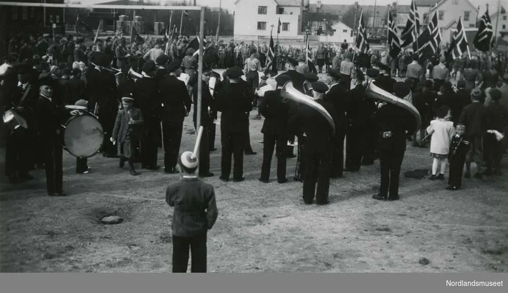 17. mai 1949 i Bodø. Folkemengde samlet på den gamle idrettsplassen. Hornmusikk og norske flagg. Bebyggelse i bakgrunnen.