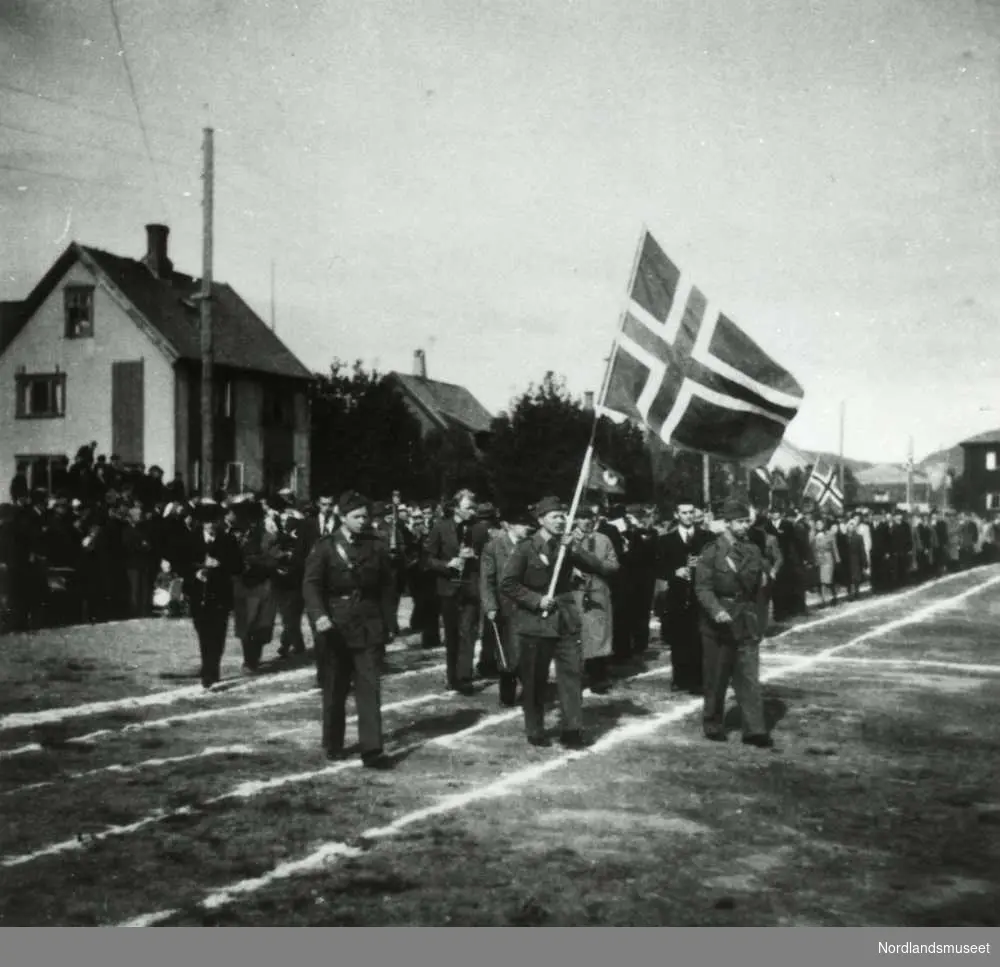 Fra 2. verdenskrig. Frigjøring. Parade for kronprins Olav i 1945. Folk paraderer på en stadion, og vi ser en del boliger i bakgrunnen.
