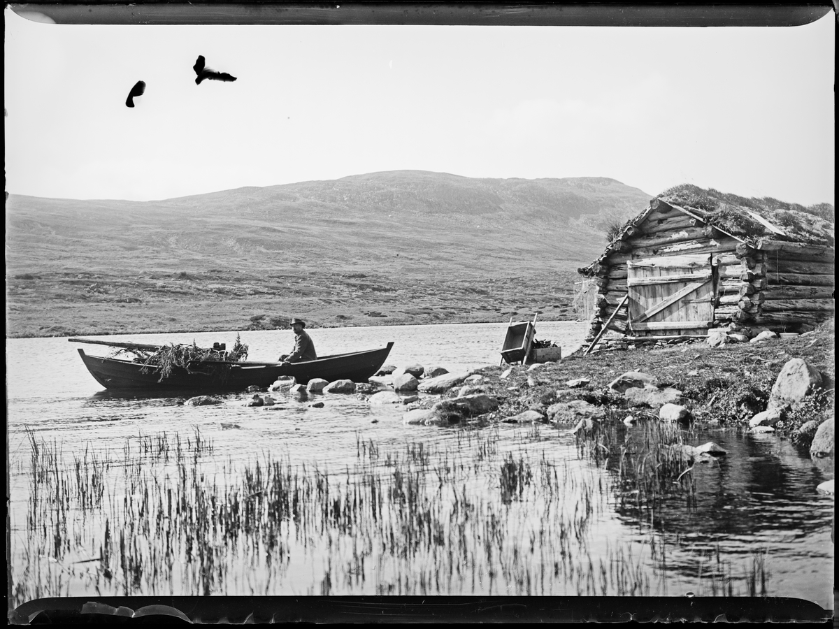 "Heimdalen 1906" (inskr eske). Fjellandskap. Sjølisætra ved Atnsjøen i Rondane. En mann sitter i en robåt. Et laftet båthus ligger rett ved vannet.