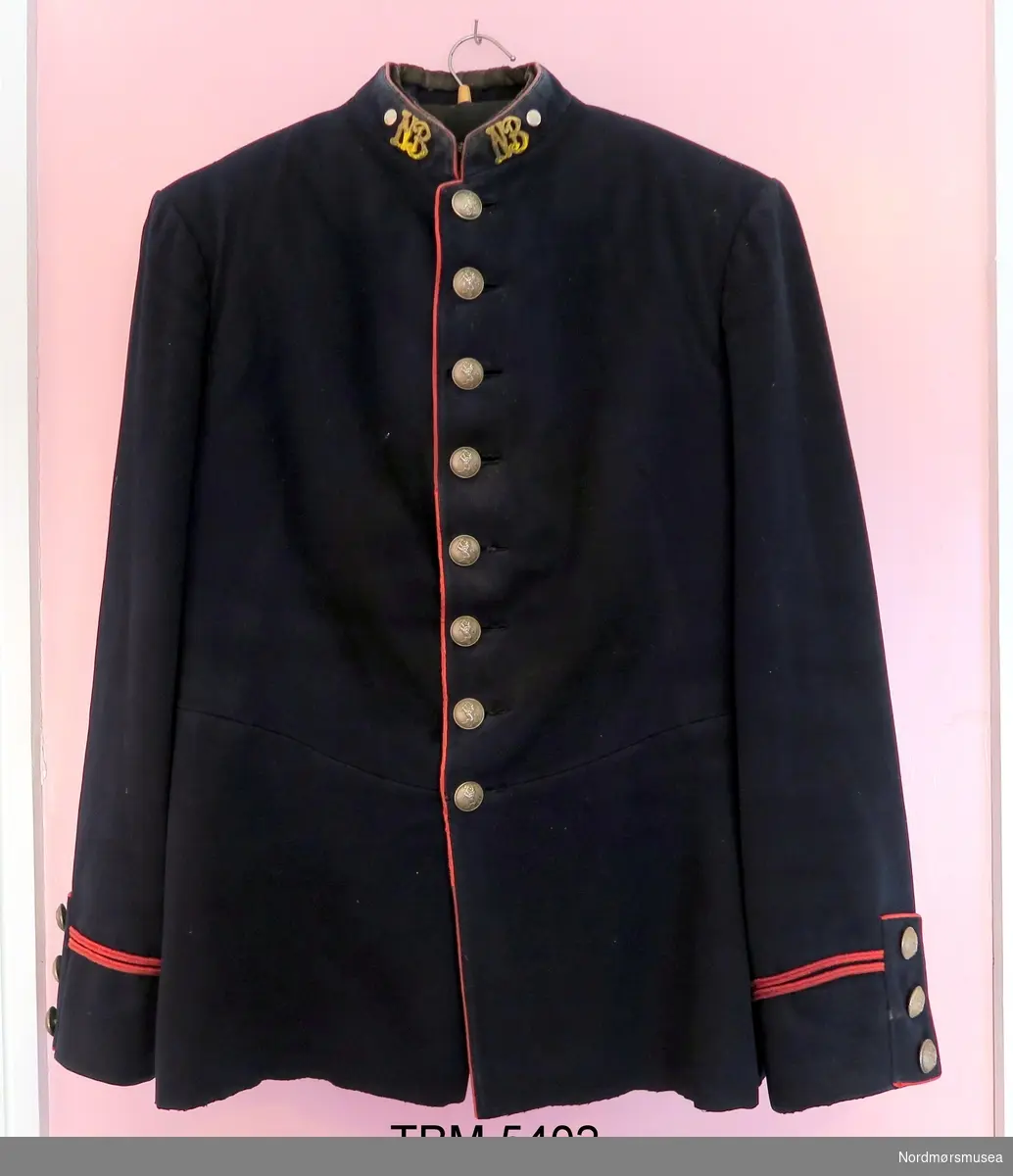 Militær jakke som er sort med raude kantar. Blanke knappar med norske løve på. På jakkekragen er det metallbokstavar, NB. Splitt med knappar og raude kantar bak.