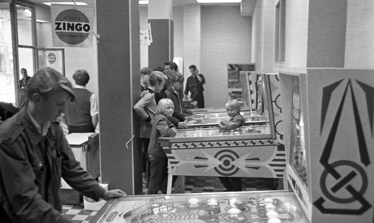 Spelautomat, 19 augusti 1965

I förgrunden står en yngling vid en spelautomat i en spelhall. I bakgrunden står flera barn och ytterligare ungdomar vid flera spelautomater. På en av pelarna i lokalen skymtar en reklamskylt med texten " Apelsin Zingo försvinnande god."