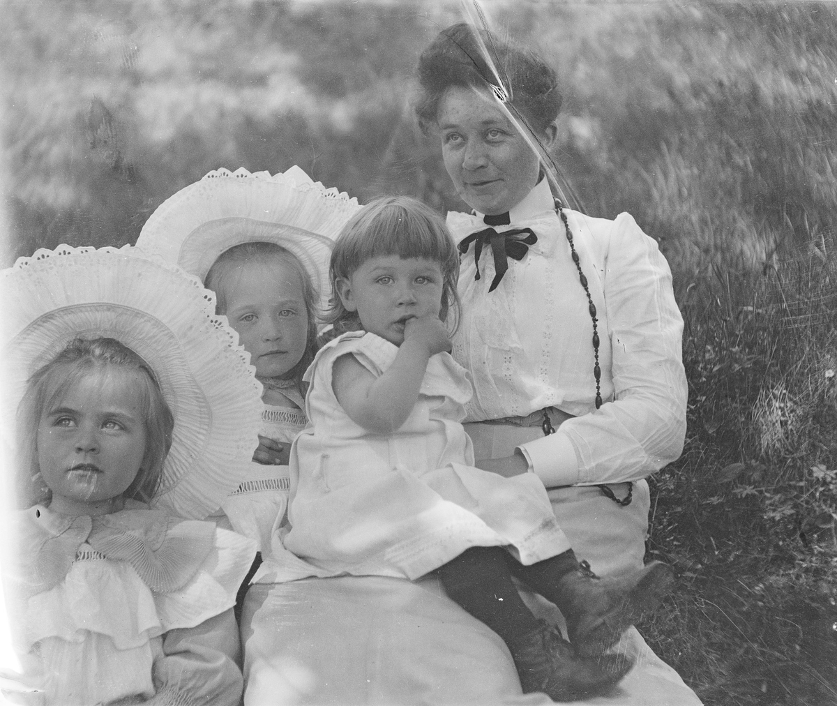 En kvinne sitter sammen med tre små barn i gresset. Den yngste sitter på fanget hennes.
