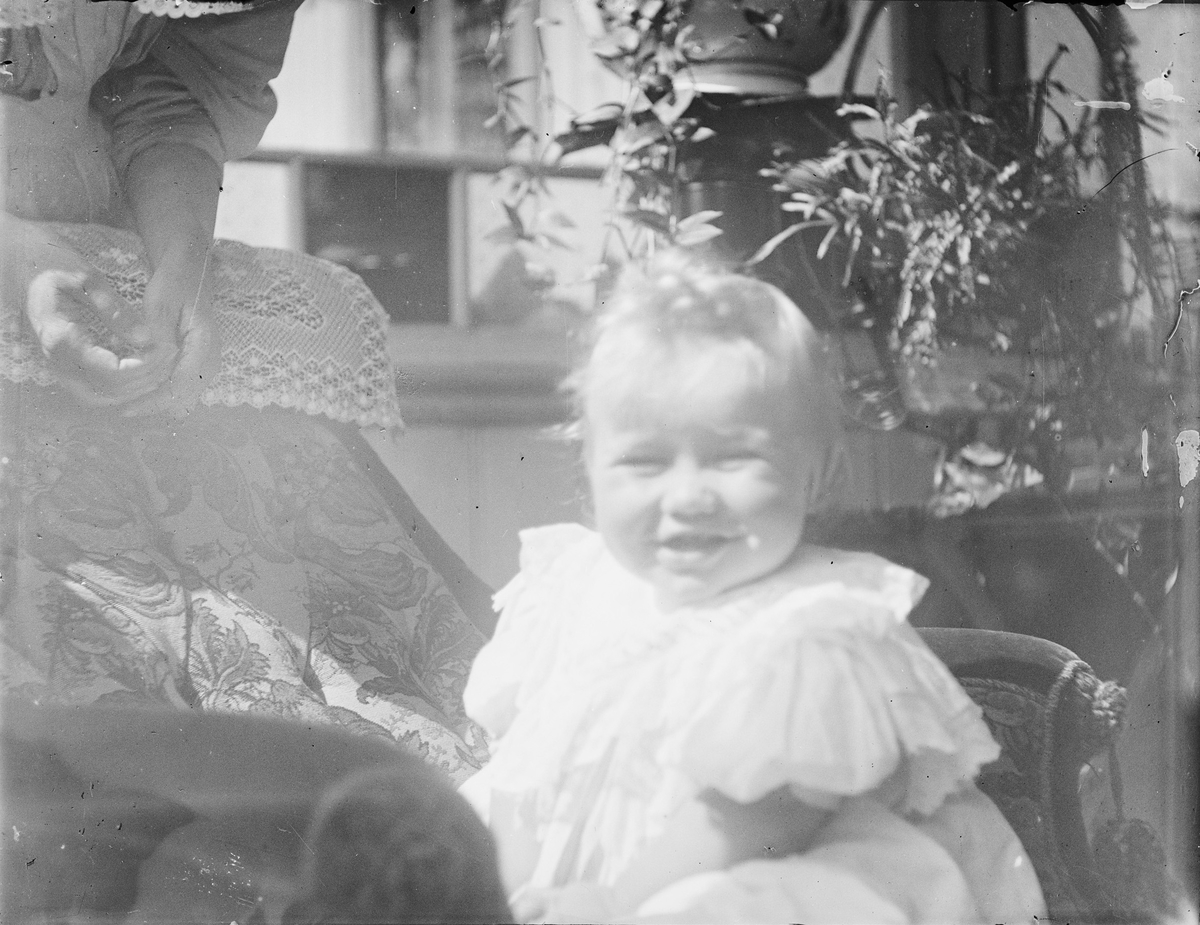 Et smilende barn med sol i ansiktet sitter i en lenestol på en innglasset veranda. I bakgrunnen ser vi hendene til en kvinne.
