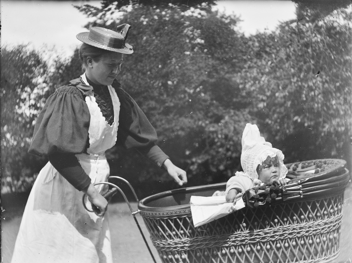 En kvinne med hvitt forkle passer på et lite barn i en barnevogn.