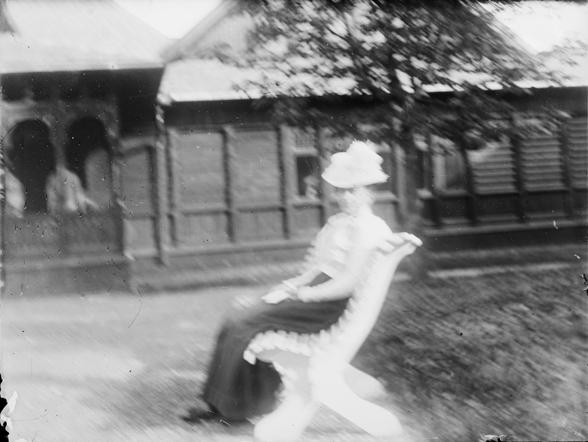 En dame sitter på en hagebenk utenfor et hus bygget i sveitser-/ dragestil. Benken står på en gruslagt plass i en opparbeidet hage. Under husets svalgang sees to andre personer.
