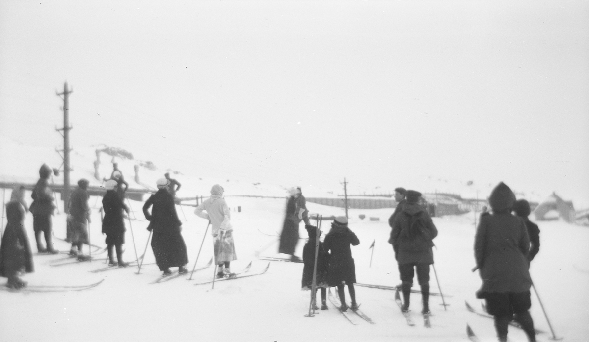 Menn og kvinner står på hver sin side av en bakke eller løype, de fleste med ski på bena. I bakgrunnen sees bygninger, fjell og en overbygget del av jernbanen.