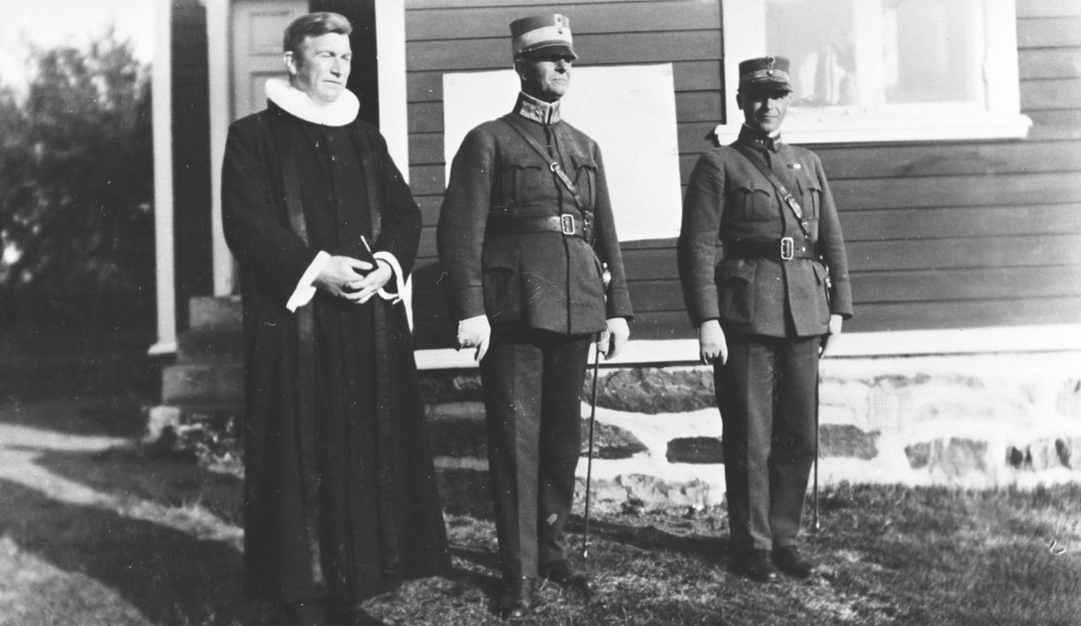 Vi antar at bildene er tatt i forbindelse med gjenopprettelse av Varanger batlajon 01.07.1934 .Bildet viser tre menn, Prost Skauge, General Jakobsen og hans adjutant i uniform foran soldaterhjemmet på Nybergmoen 1934. Skauge holder hendene rundt en bibel.