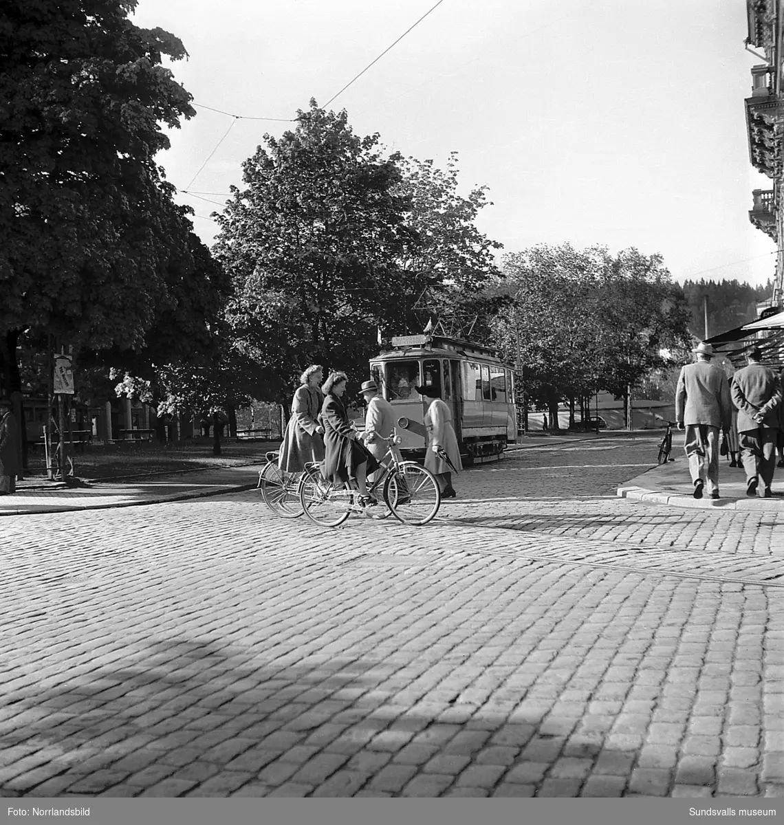 Cyklister och flanerande människor på Esplanaden, vid korsningen Storgatan, och bakom närmar sig spårvagnen på väg mot Kubikenborg.