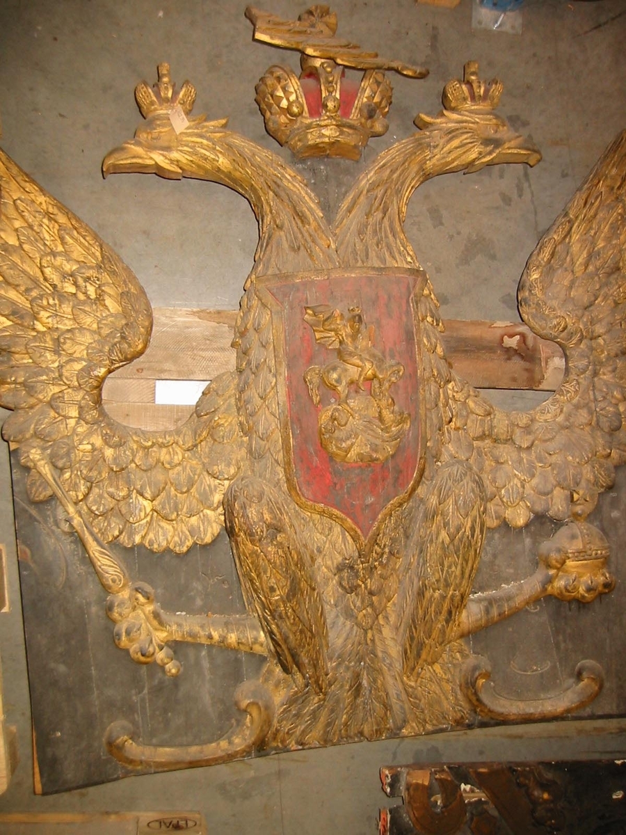 Akterspegelsornament. Ryska tsar- (kejsar-?) dömets vapen: krönt, förgylld dubbelörn med röd sköld, varpå är fästad en skulptur av St. Göran och draken.
Sjöfynd, restaurerat och kompletterat.