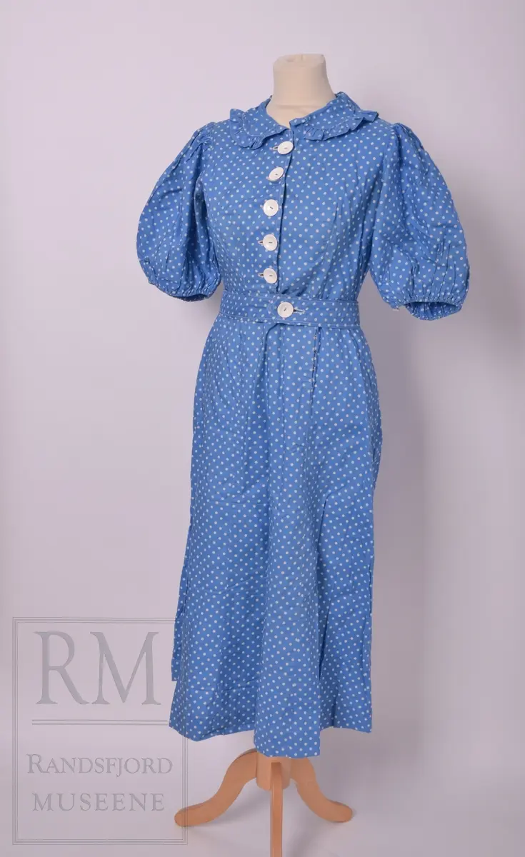 Sommerkjole m/hatt. a) Bomullstoff, blått med hvite prikker. Sammensydd i livet. Overdelen har Lukning foran med 5 hvite knapper og knapphull, puffermer. Krage med heppekant. 6 Bredder i utskrådd skjørt. Lukning til venstre for midt foran. 
b) Belte med knapp og knapphull. 
c) Hatt, Heklet av hvitt bomullsgarn. Ståltråd ytterst i Bremmen (rusten) Figurkant på pullen. Pyntebånd av kjoletøyet.