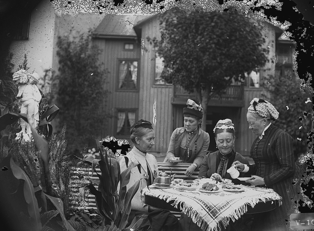 Kaffedrickande, fyra kvinnor vid kaffebordet i trädgården.
Bostadshus i bakgrunden.