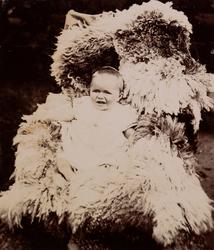 En baby (trolig Erica Mathiesen) sitter på et saueskinn i en