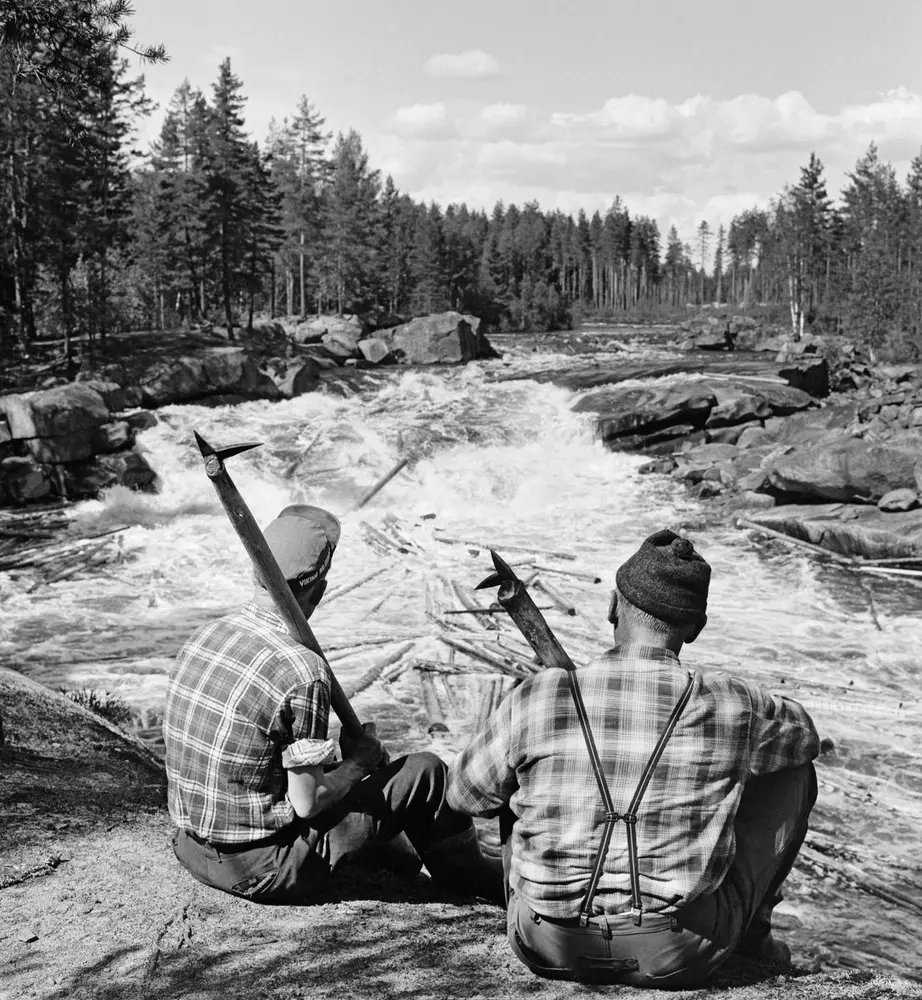 To tømmerfløtere, utstyrt med fløterhaker og kledd i rutete bomullsskjorter, fotografert sittende på en bergnabb nedenfor en foss i den øvre delen av Flisavassdraget