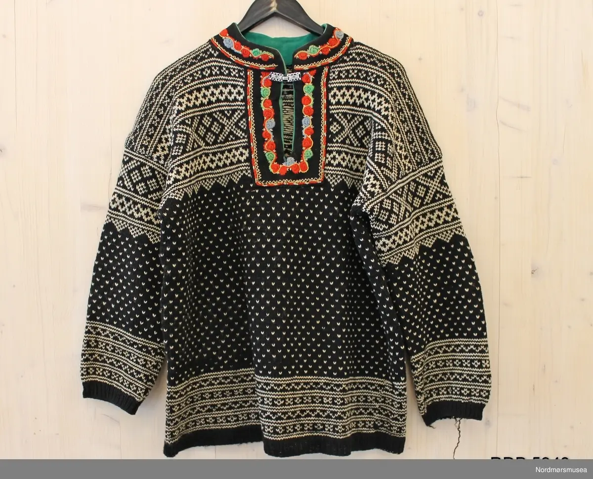 Håndstrikket Setesdalsgenser, herregenser, svart men kvitt mønster og med påsydd brodering i halsen. Broderiet har grønt fôr, og en tinnhekte påsydd foran. Genseren er slitt og hullet på albuene.