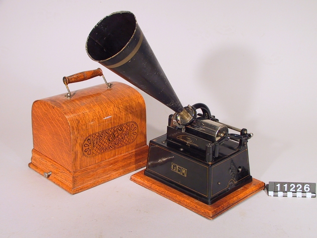 Fonograf, enligt Edison. Mindre typ, fjäderverksdriven, jämte "recorder" för upptagning. Recorderns nr. 10882. Monterad på träplatta. Skyddshuv med handtag.