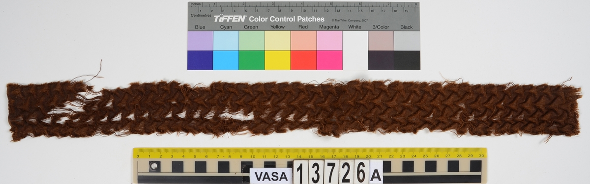 Textil. 
Fyndnummer 13726 är uppdelat i a och b.
Fnr 13726a är ett mönstervävt band med invävda/insydda metalltrådar. År 1983 och vid dokumentering 2015 syns dock endast hålen efter dessa. 
Fnr 13726b är ett silkesband vars ändar är knutna till en rosett.