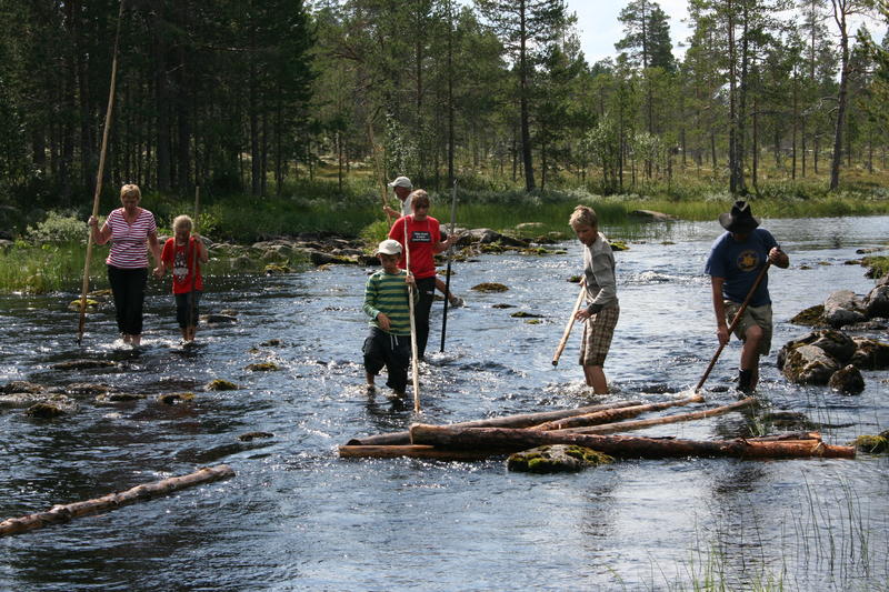 A group of children and adults testing out log driving. They are walking in the river with hooks to sort the timber that floats. .