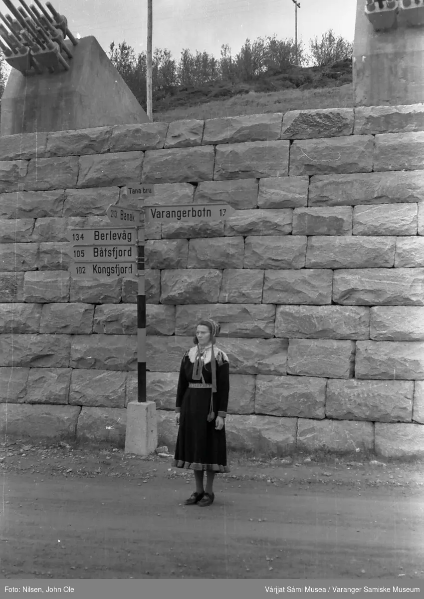 Signe Nilsen står ved brohodet på østsiden av Tana Bru. Bak henne står det et skilt som viser distansen til ulike steder i Øst-Finnmark. 26. juni 1966.