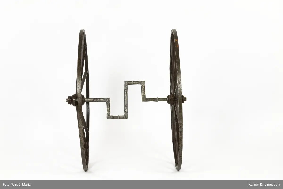 KLM 12224:1-5. Velociped, trehjuling. Fem delar. Av hemgjord typ. Flera delar till ramen saknas. Tre stycken hjul av trä, målade i brunt med gul dekorrand. Hjulen är järnskodda och ett av de stora hjulen har stämplar i järnet: 1868 samt EA inom en cirkel. Mellan de två stora hjulen (:1-:2) sitter en axel av metall (:3) för trampor, trissor, sannolikt av trä, som kunde snurra runt. Det minsta hjulet (:4) har fungerat som fram- eller bakhjul. På hjulet är en del av ramen monterad. Längst upp ett styrstagsliknande tvärstag (:5) med ett litet runt genomgående hål i respektive ände för infästning, i detta fästes sannolikt det som reglerade styrningen. Själva tvärstaget har inte använts som styre.