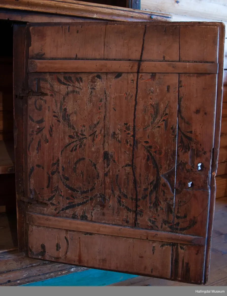 Rester etter rød og blå maling.  Gesims, skråner ned bakover. Profilert. 6 sirkulære hull i arkitraven.  Mellom disse er malt med sorte bokstaver.  Gavlene har utskårne "hylleknekter".  Bakveggen består av fire vertikale planker.  Skjøtene er dekket med profilerte lister.  Bare to biter i nederste del til venstre er fremdeles på plass.  Nederste del er lukket skap.  Tredelt, midterste del er skapdør.  Platen har profillist.  Feltene har speilfyllinger.  Blåmalte.  Klosset marmorering.  Speilfyllingenes form er som en liggen H.  Har profillist.  Skapet er satt på en ny grønmalt plate.  På døren er malt hvite bokstaver.  Nederst på døren er skåret inn bokstaver, opp-ned.  To nøkkelhull m. jernbeslag på venstre side av døren.  Over døren til venstre, en dreibar trekloss, holder igjen.  Innvendig har døren merke etter stort jernbeslag på samme side som nøkkelhullene.  To lister skåret i treverket minner om lange jernbeslag,-smalner mot nøkkelhullene.  Akantusranker i rødt, blått og hvitt, meget avslitt.  En hylle i skapet. Mangler bakvegg.  Karmen har på venstre side spor etter to jernhengsler.  Øverst på høyre kortside, mangler gesimsen en bit, og holdes til sideveggen med en jernkrampe.  Jernkrampe også på venstre side.  Skapplaten mangler list på høyre side.  Venstre side, fra gesimsen og ned har spor etter å ha stått inntil en vegg eller et annet møbel.