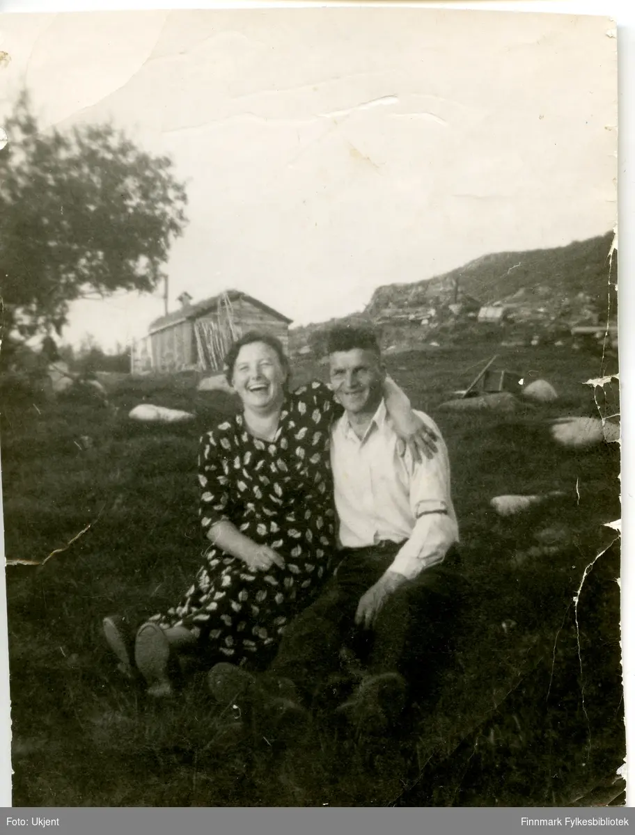 Et portrett av ekteparet Johanna og Petter Aule. Johanna er kledd i kjole, Petter i skjorte og bukse. De sitter i gresset på bakken, og i bakgrunnen kan man se en bygning laget av tre.