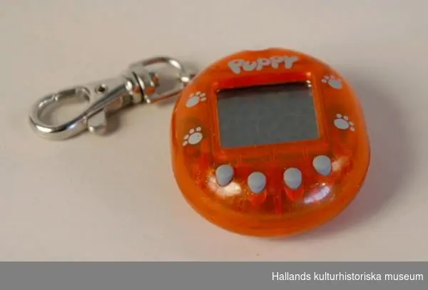 Elektronisk leksak, tamagotchi. Orange plasthölje kring en liten bildskärm, metallkedja med hake för upphängning. Text: "PUPPY".