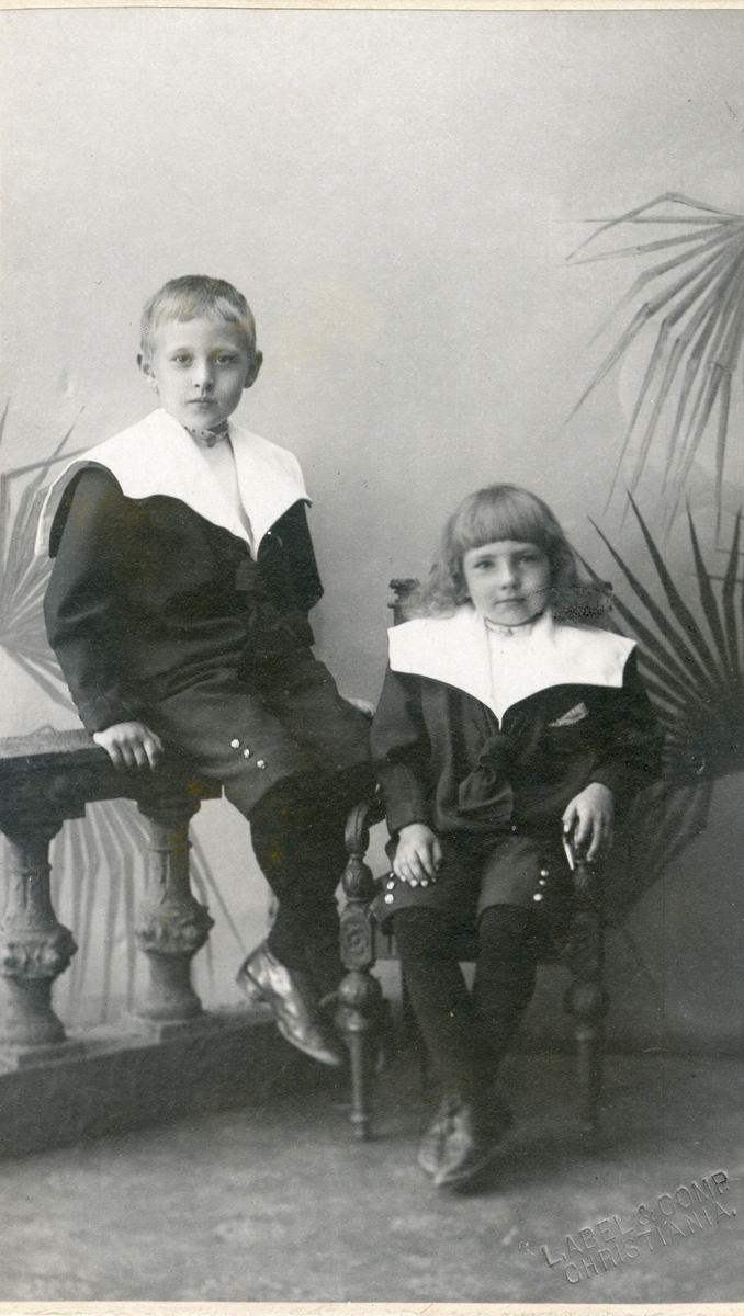 To barn avbildet sittende på en stol og gelender. Begge er iført jakker med store slag.