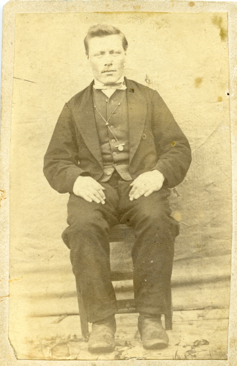 Portrett av en mann sittende på en stol foran et lerret. Mannen er iført dress med vest og skjorte.