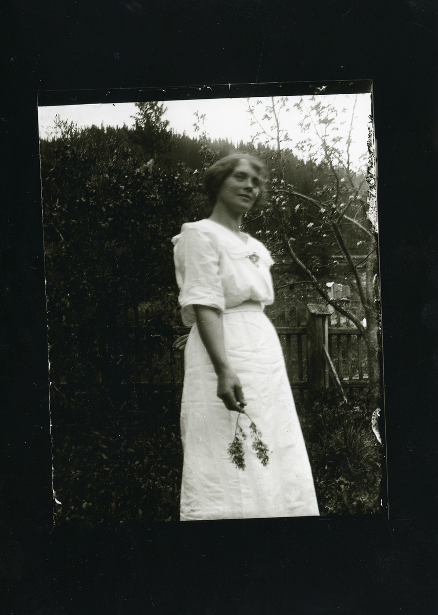 Kvinne iført en hvit kjole med en blomst i hånden. Kvinnen står foran en hekk og en port.