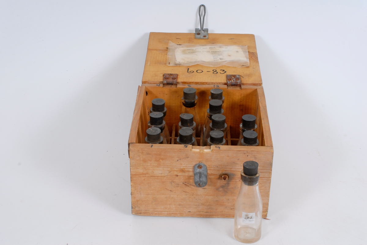 Form: tilnærmet kvadratisk kasse med 13 små glassflasker i, flaskene har gummikork og nr.

